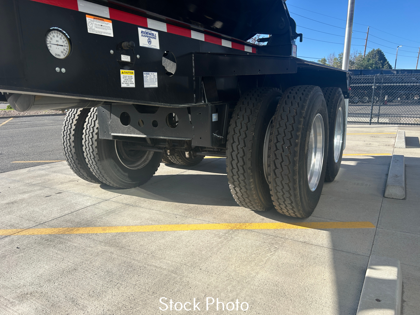 Truck trailer rear dual-axle, black frame with warning labels, steel wheels, see-through ruler marks, parking lot; stock photo of a new Smithco SX2-4034 dump trailer.