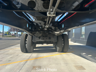 Underbelly view of a dump trailer showing dual rear axles, suspension, and chassis components; cables and metal framework visible; stock photo label on ground.