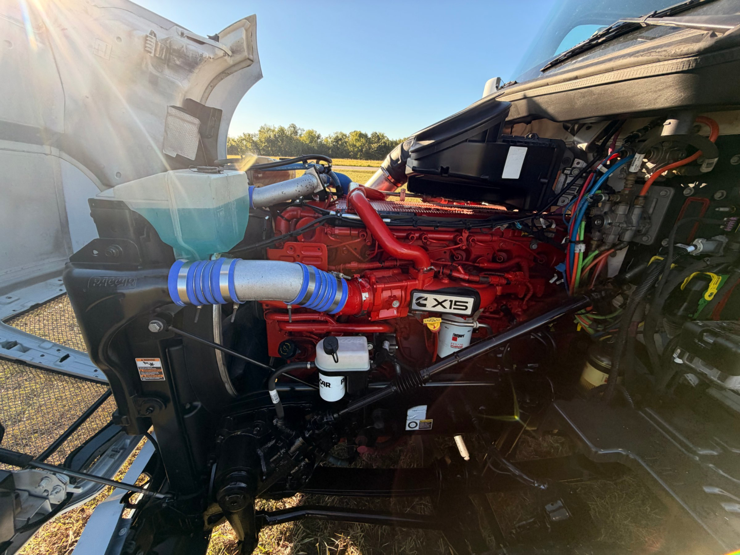 Kenworth T680 engine bay showing a red engine with blue hoses, multiple belts, and various components in the right-hand drive cabin area.