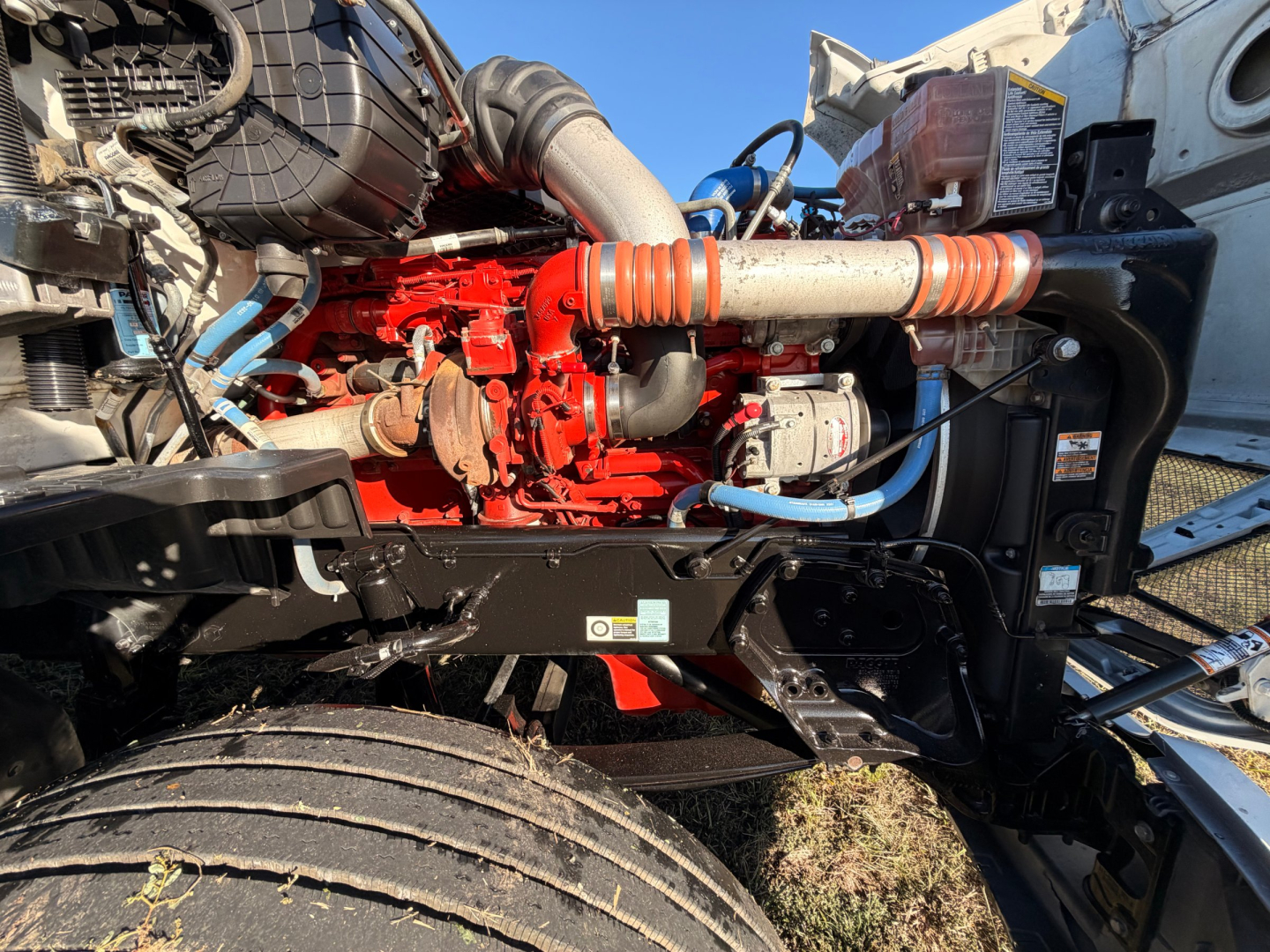 Kenworth T680 engine bay side showing red engine block with blue hoses, black frame, large intake hose, and orange coolant hose. Visible labels and components but no legible text.