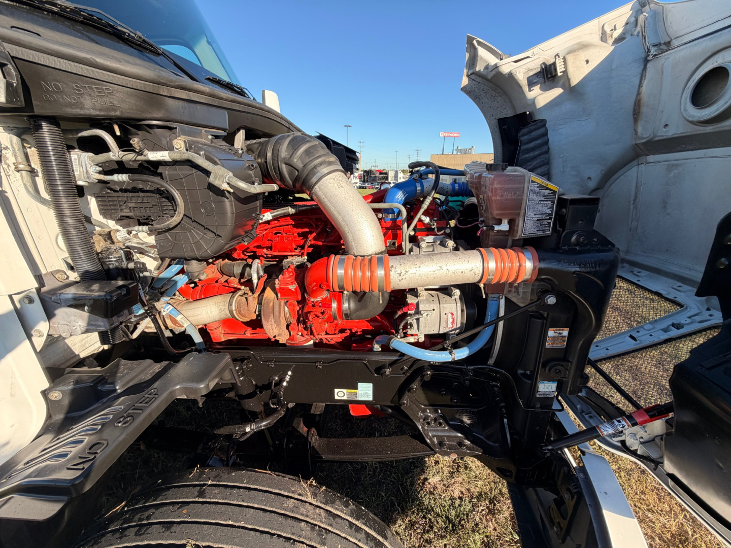 Kenworth T680 engine bay with red engine components and intake hoses visible; tags, hoses, and metal framework in view.
