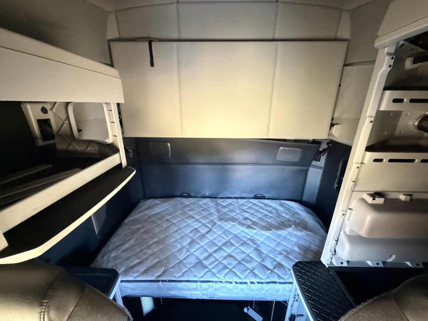 Cabin bunk area of a 2022 Kenworth T680 sleeper truck showing a bed with quilted mattress, overhead storage shelves, and side panels.