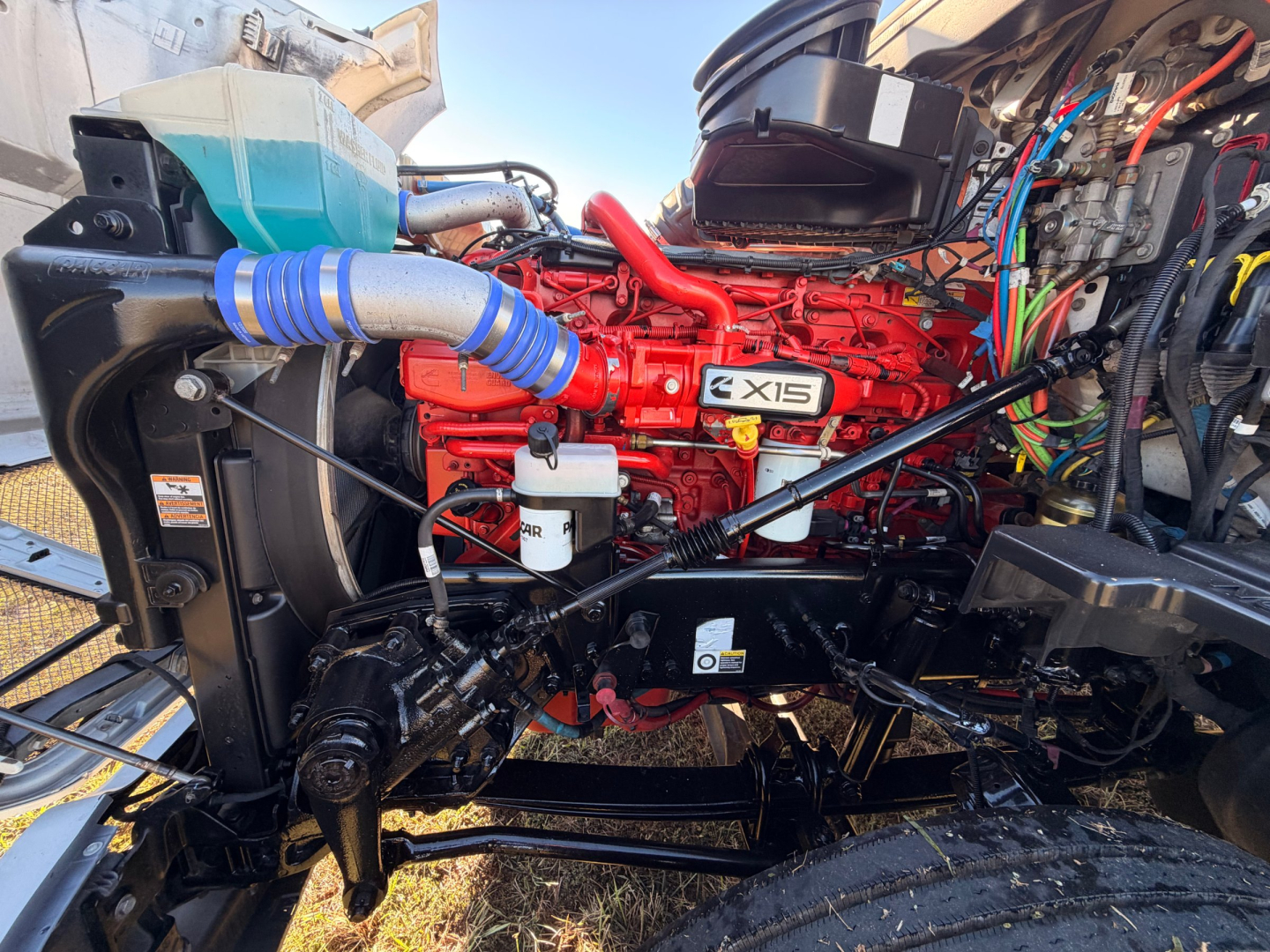 Kenworth T680 engine bay showing red X15 engine, multiple hoses, wiring, hydraulic lines, and front suspension components.