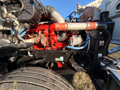 Kenworth T680 engine bay side showing red engine block with blue hoses, black frame, large intake hose, and orange coolant hose. Visible labels and components but no legible text.