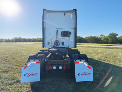 Trailerless Kenworth T680 rear view showing fifth wheel and dual rear axles, white mud flaps with MHC logo, open cab area, clear sky.