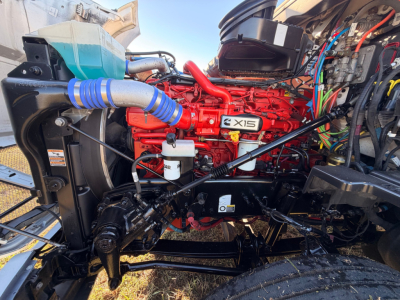 Kenworth T680 engine bay showing red X15 engine, multiple hoses, wiring, hydraulic lines, and front suspension components.
