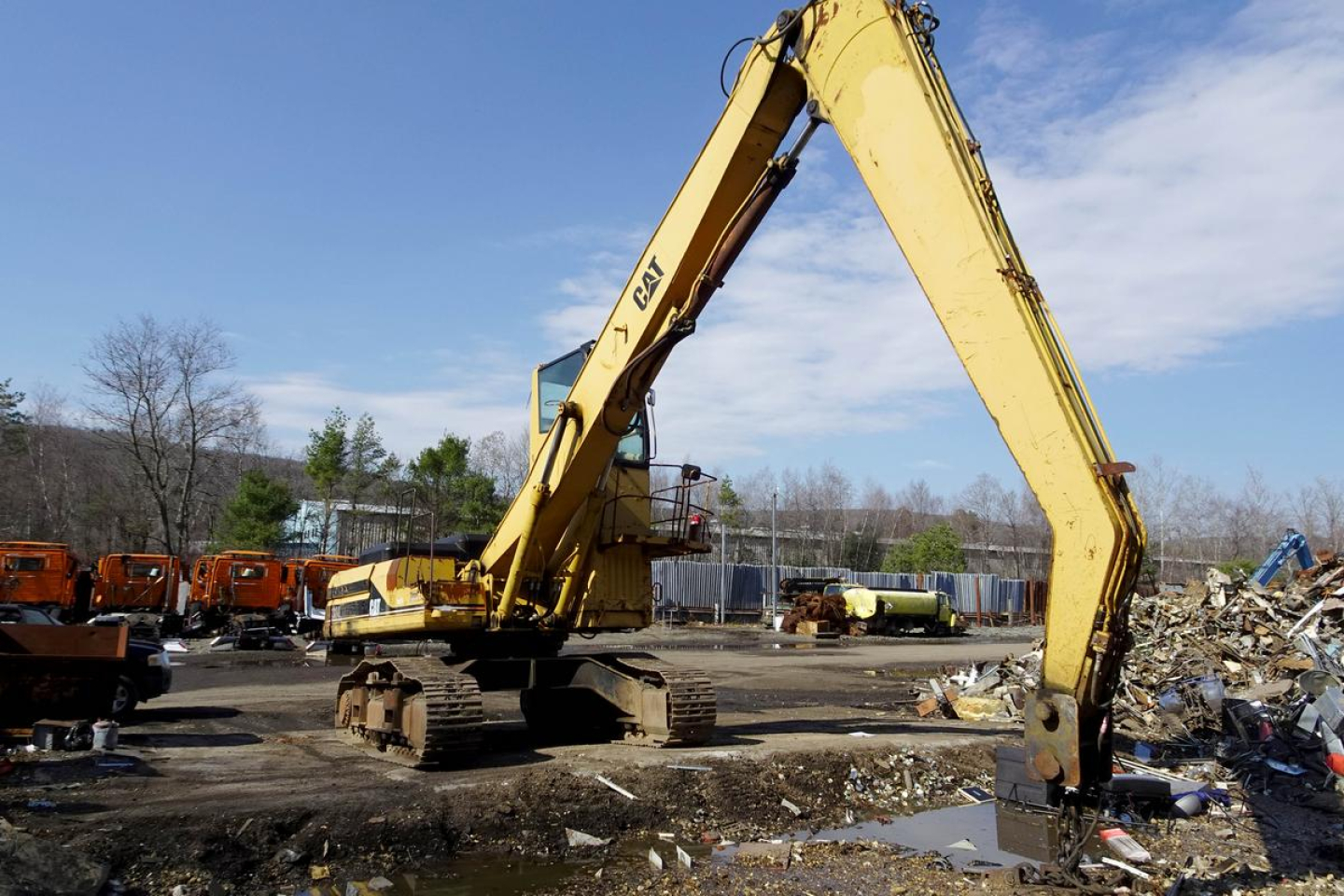 Caterpillar 330L MH excavator in a salvage yard with extended arm and bucket, yellow paint, tracked undercarriage, piles of debris and other heavy equipment in background.