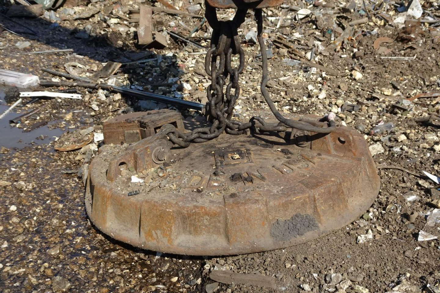 Circular metal Caterpillar 330L MH undercarriage weight plate with chains attached, resting on gravel and debris.