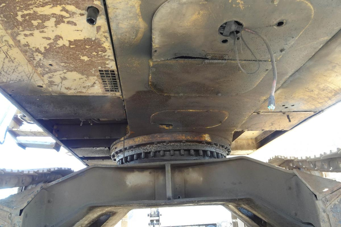 Underbelly view of a Caterpillar 330L MH tracked vehicle showing the rotating undercarriage and wear on the hull; mechanical coupling and debris visible.
