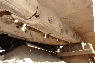 Tracked undercarriage track links with pins and rusty metal surfaces; dirt and grime on track and undercarriage components.