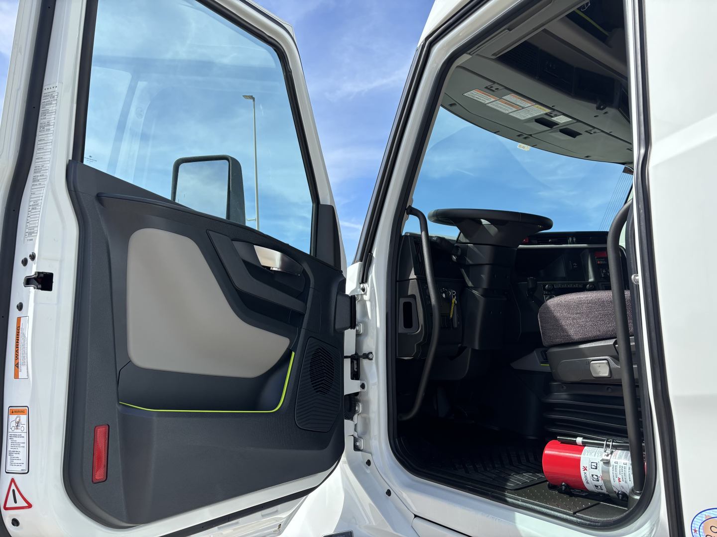Volvo VNL cab interior and open door showing driver seat and steering wheel with dashboard, entry area, and a fire extinguisher on the floor.