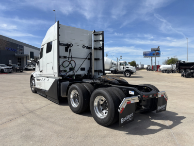 Volvo VNL cab and chassis viewed from rear-left, showing rear axles, fifth-wheel area, and side mirrors with fuel tanks. Clear blue sky and dealership lot in background.