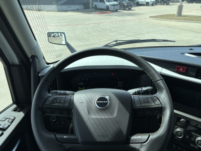 Volvo truck cabin steering wheel and dashboard view in a new 2027 Volvo VNL64T640 sleeper with Volvo emblem on the airbag cover and control buttons on both sides of the wheel.