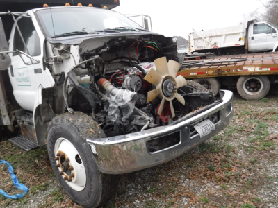 Front view of a damaged 2011 Ford F-750 dump truck with exposed engine and radiator, large cooling fan, front bumper chrome, and flatbed trailer in background.