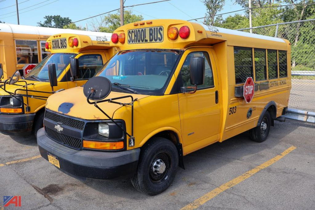 Yellow Chevrolet Express school bus in a parking lot showing front view with stop sign, school bus lettering, and rooftop 'SCHOOL BUS' sign.