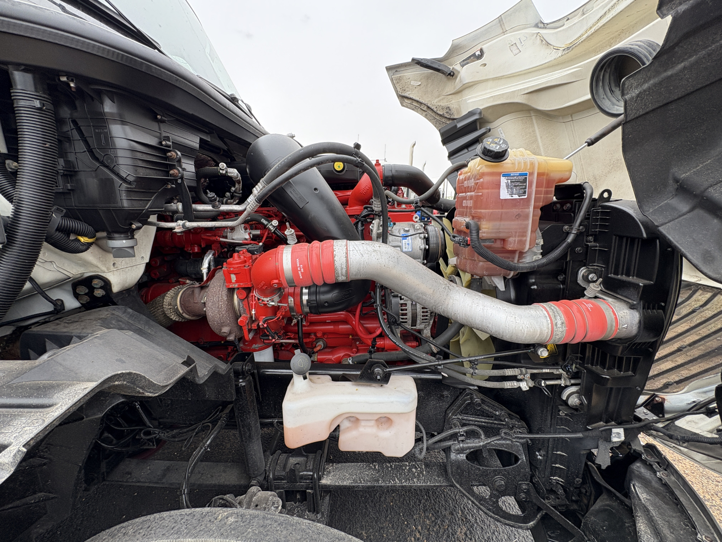 Truck engine bay showing red engine block, hoses, coolant reservoir, and various fluid lines in a Daycab configuration.