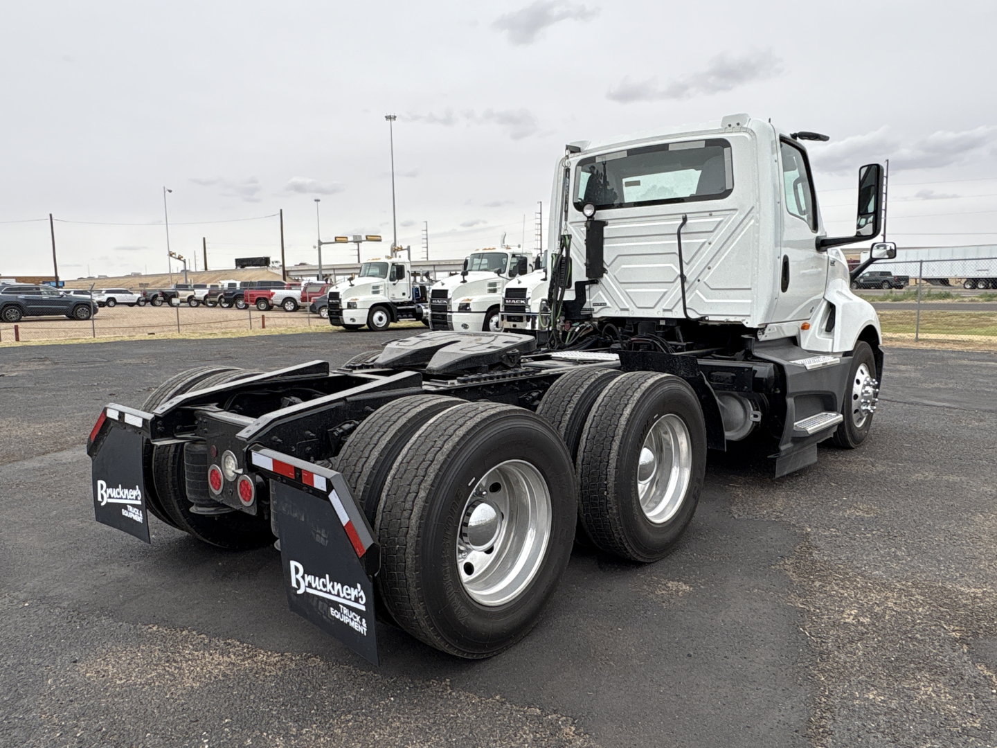 Truck rear/side view showing chassis and dual rear axles, with mudflaps labeled Buuckney? Buckneck? hardware. Visible text on mudflaps: Brackets? The image includes a white International LT625 daycab tractor in a lot.