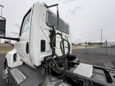 Truck cab of a 2021 International LT625 conventional daycab, side view showing roof-mounted mirrors, rear cab area with green hoses, metal diamond-plate steps, and hitch area.