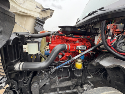 Truck engine bay showing red Cummins ISX engine and black intake hoses in a daycab, with yellow coolant reservoir and various wiring harnesses.