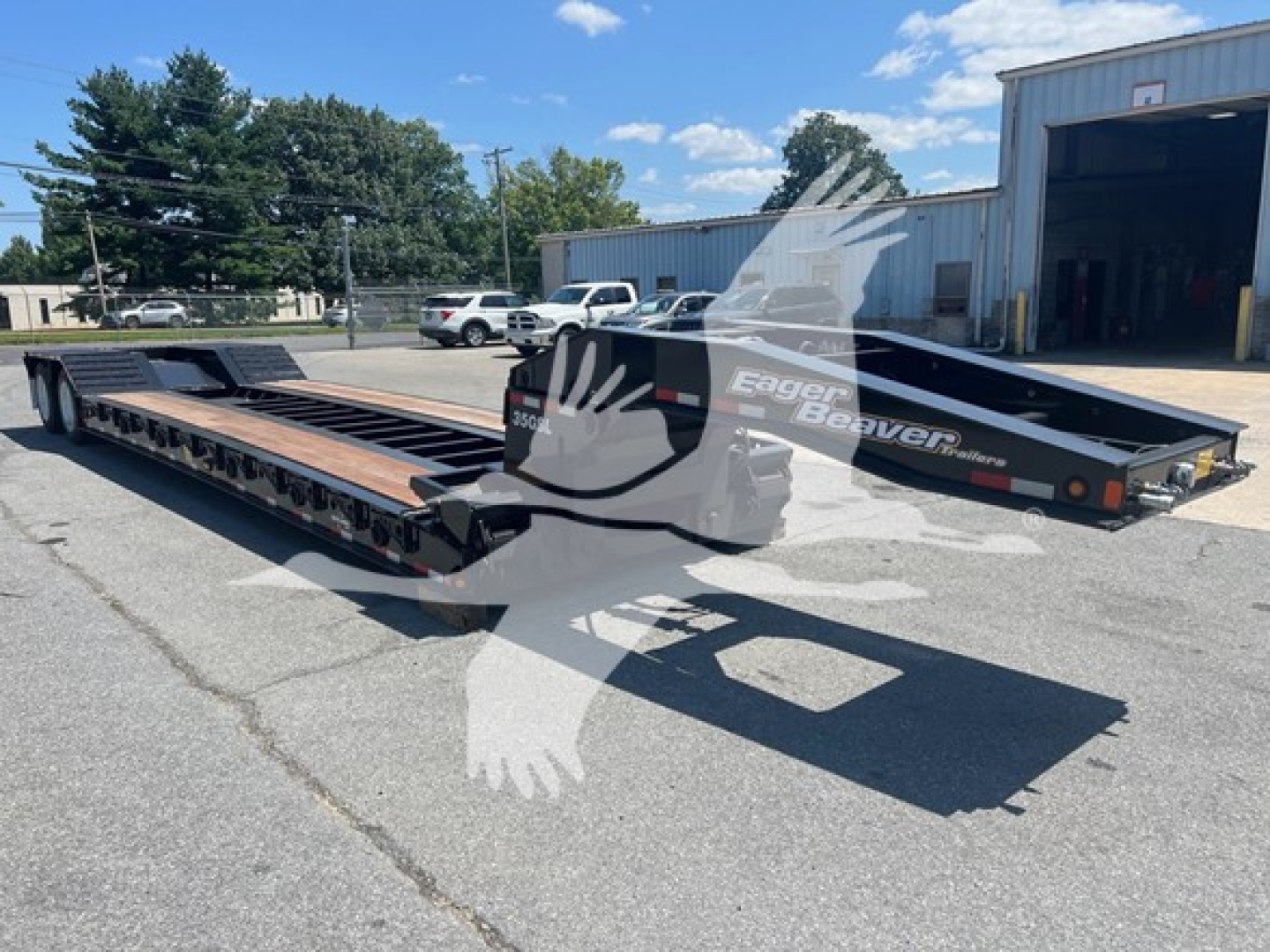 Lowboy trailer in a parking lot showing the long flatbed with black frame and wooden deck, Eager Beaver logo on side and ramp at rear.