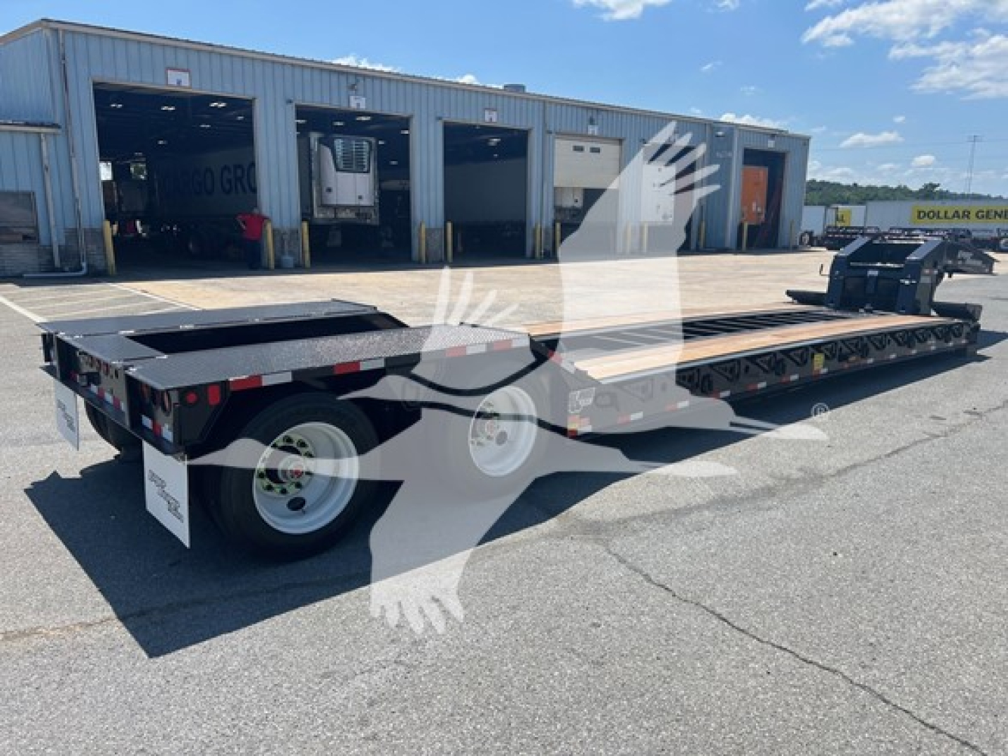 Lowboy trailer showing flatbed deck and multiple axles with dual rear wheels and ramps, black frame, warehouse background.