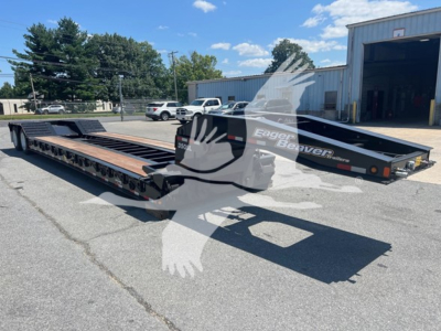 Lowboy trailer in a parking lot showing the long flatbed with black frame and wooden deck, Eager Beaver logo on side and ramp at rear.