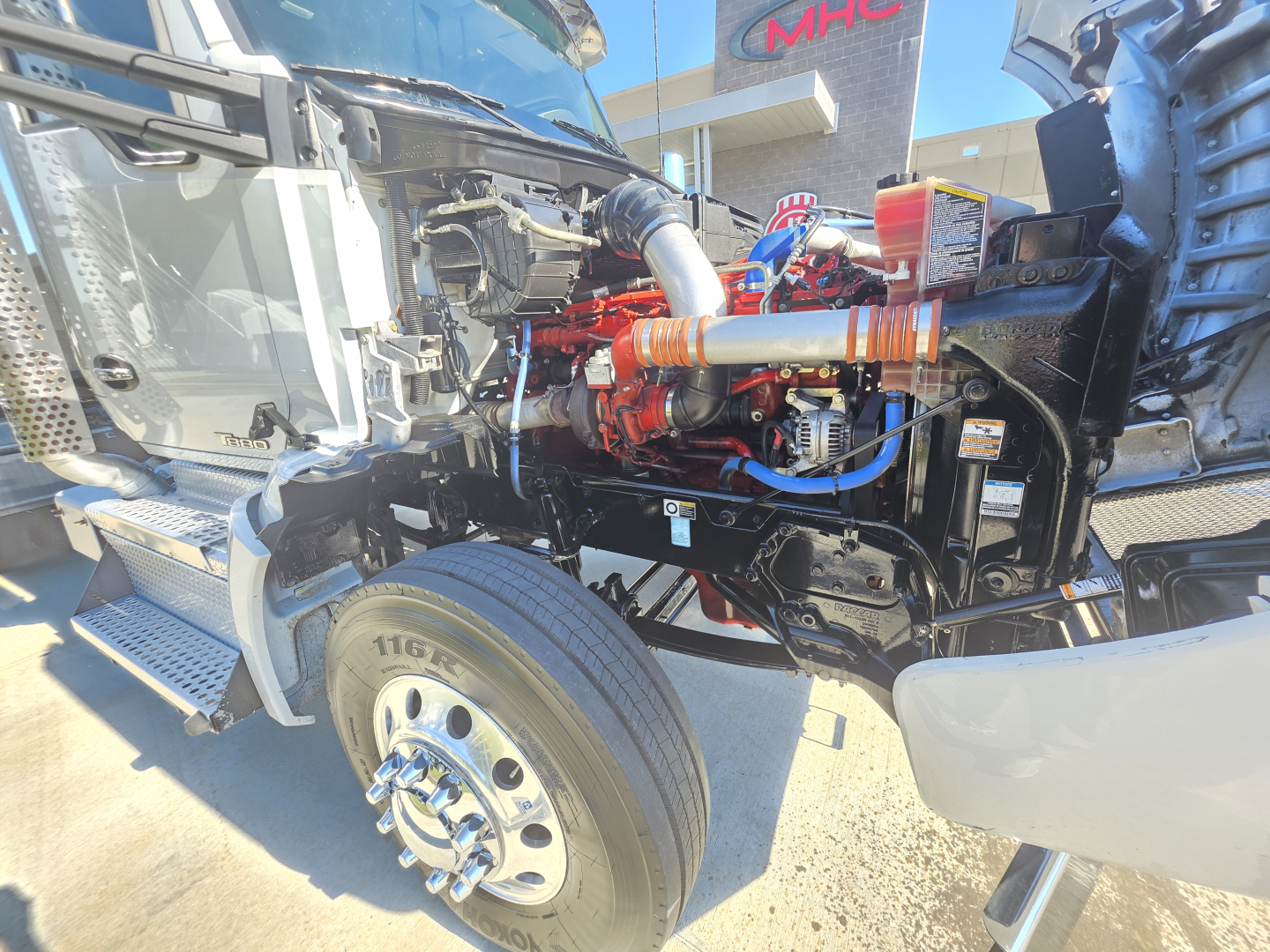 Kenworth T880 engine bay visible with red engine components, blue hoses, chrome wheel, front fender; no obvious text readable besides small labels on hoses