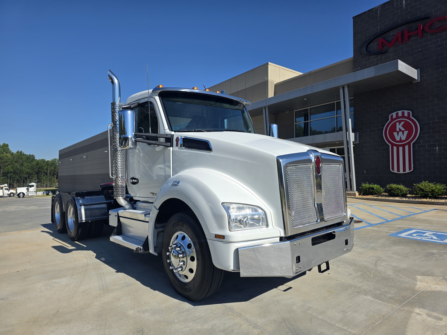 Kenworth T880 conventional daycab truck in a parking lot, chrome front grille, white cab, dual rear axles, side exhaust, KW logo visible on building