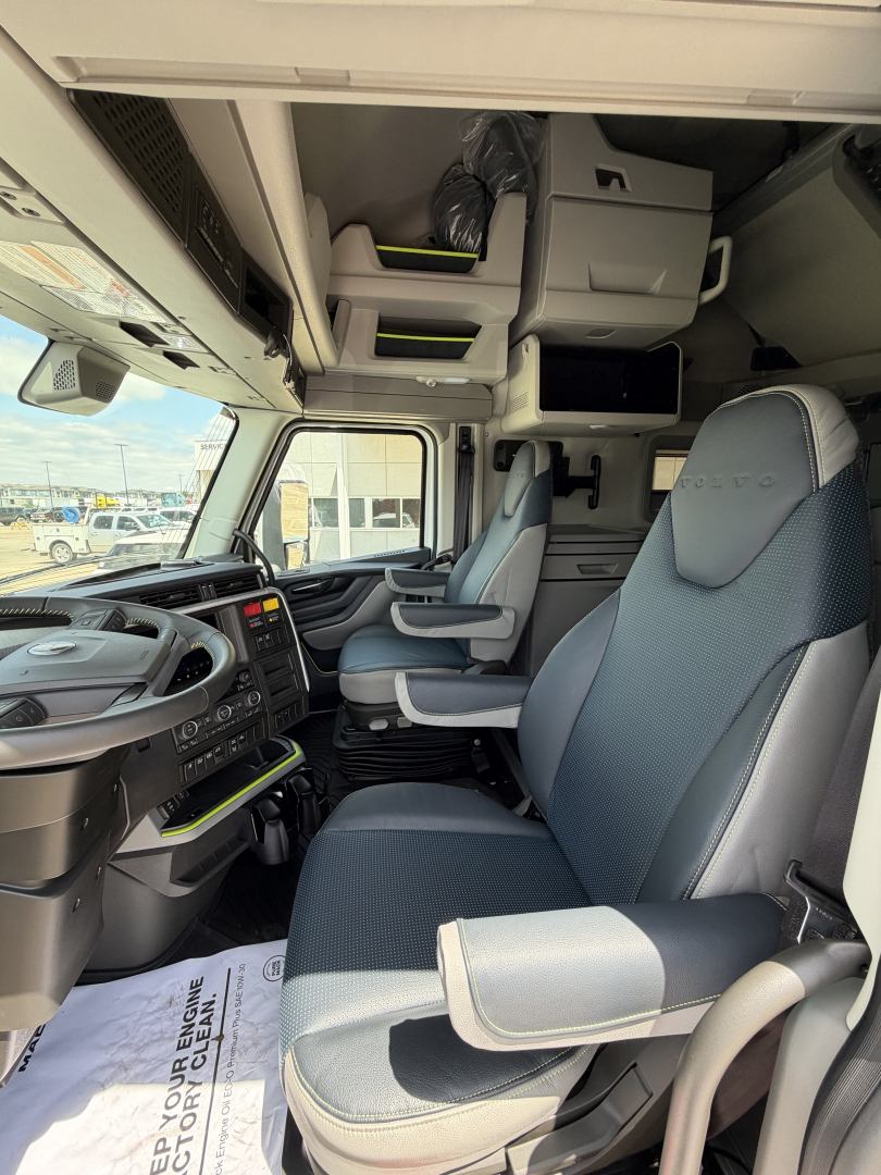 Cab interior of a Volvo truck showing captain seats, overhead storage, and dashboard with steering wheel on left; visible window and high ceiling bunk area.