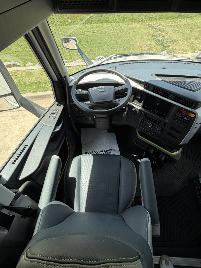 Interior view of a Volvo VNL cab showing steering wheel, dashboard, and driver seat; windshield, side mirror, and pedals visible.