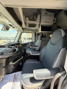 Cab interior of a Volvo truck showing captain seats, overhead storage, and dashboard with steering wheel on left; visible window and high ceiling bunk area.