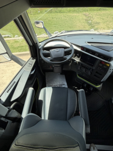 Interior view of a Volvo VNL cab showing steering wheel, dashboard, and driver seat; windshield, side mirror, and pedals visible.