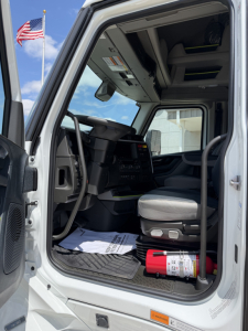 Truck cab interior of a new 2027 Volvo VNL64T860 with open door, showing steering wheel, dashboard, driver seat, and a red fire extinguisher on the floor near the doorway.