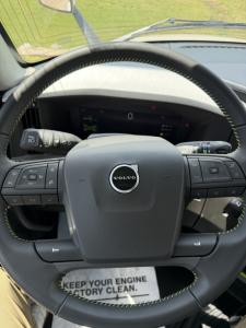 Volvo VNL64T860 steering wheel and dashboard view. Visible text on dashboard and controls includes Volvo logo on center, various steering wheel buttons, and a floor mat with the phrase “KEEP YOUR ENGINE FACTORY CLEAN.”