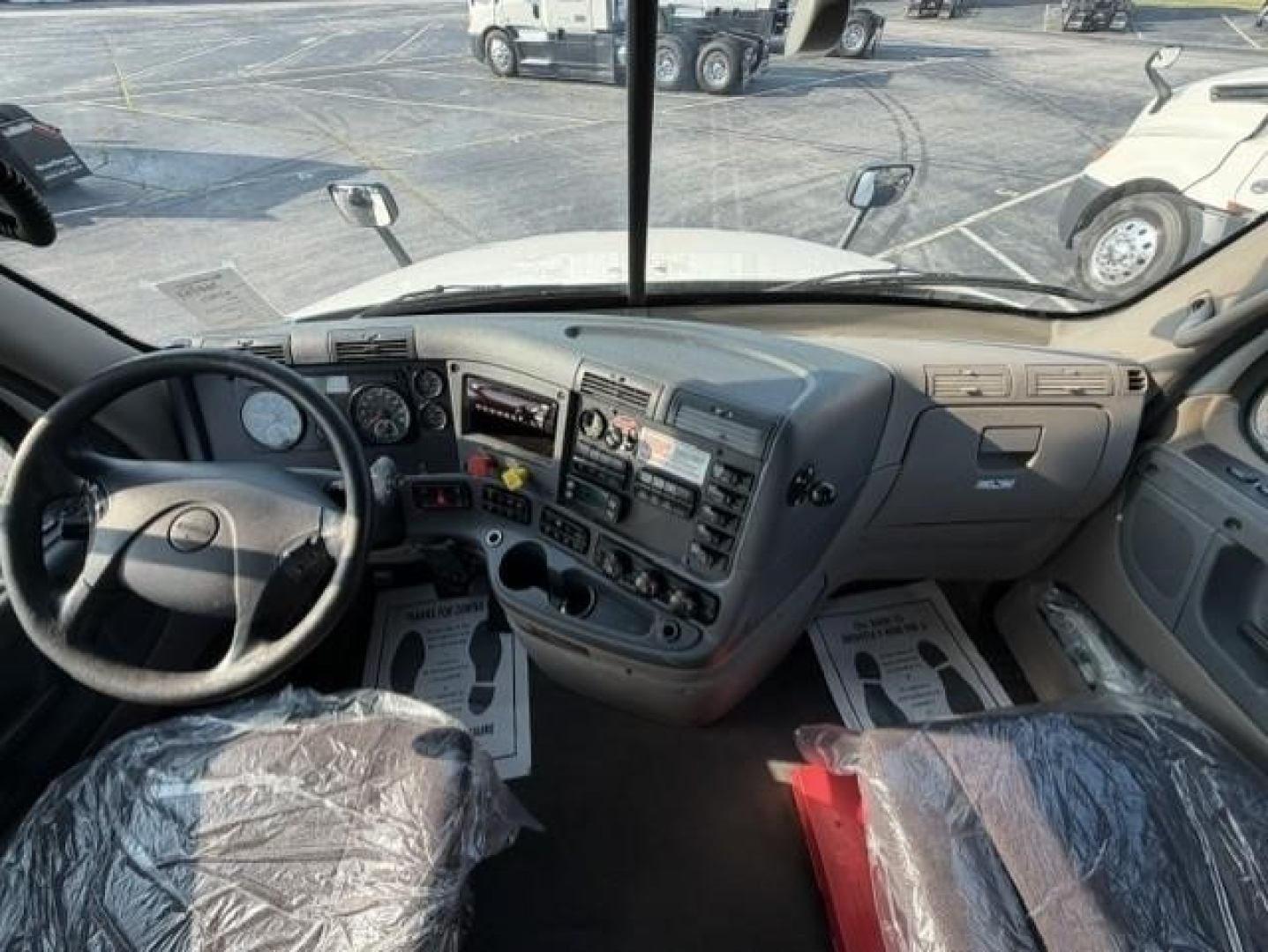 Cab interior of a Freightliner Cascadia 126 day cab showing steering wheel, dashboard, and driver and passenger seats covered in plastic.