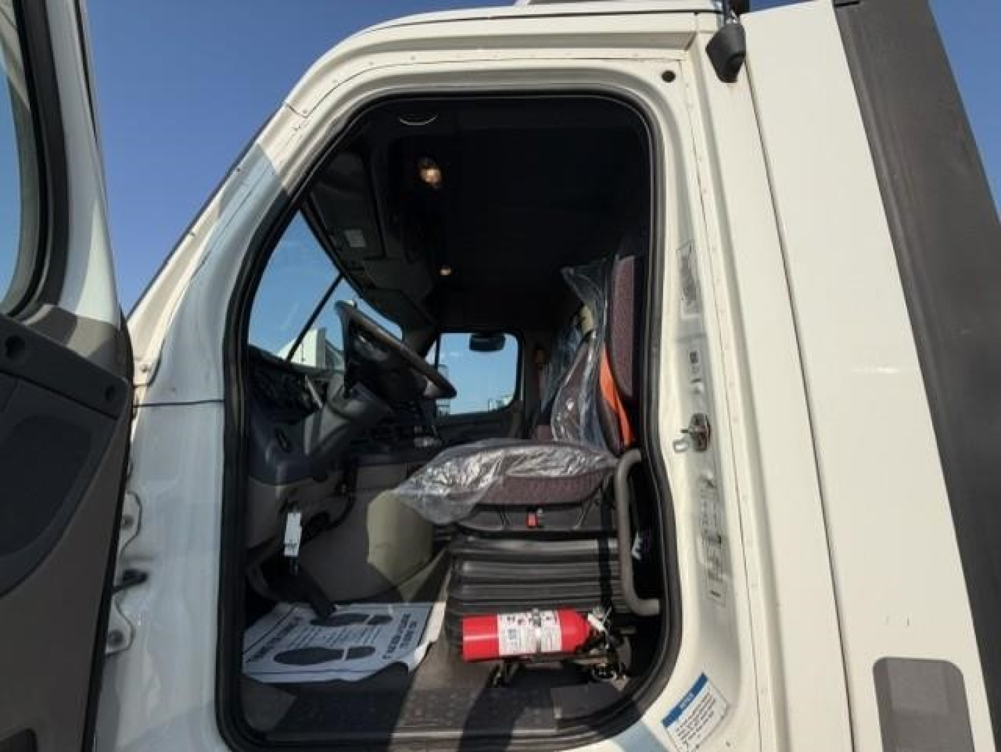 Truck cab interior with open door showing steering wheel, driver's seat wrapped in plastic, safety cone, and a fire extinguisher on the floor; windshield and dashboard visible.