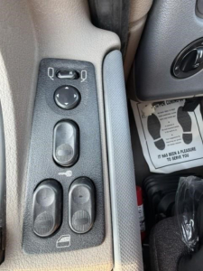 Controls in Freightliner Cascadia cab: window/mirror adjustment panel with circular joystick and two window/lock switches; partial floor mat and dash visible.