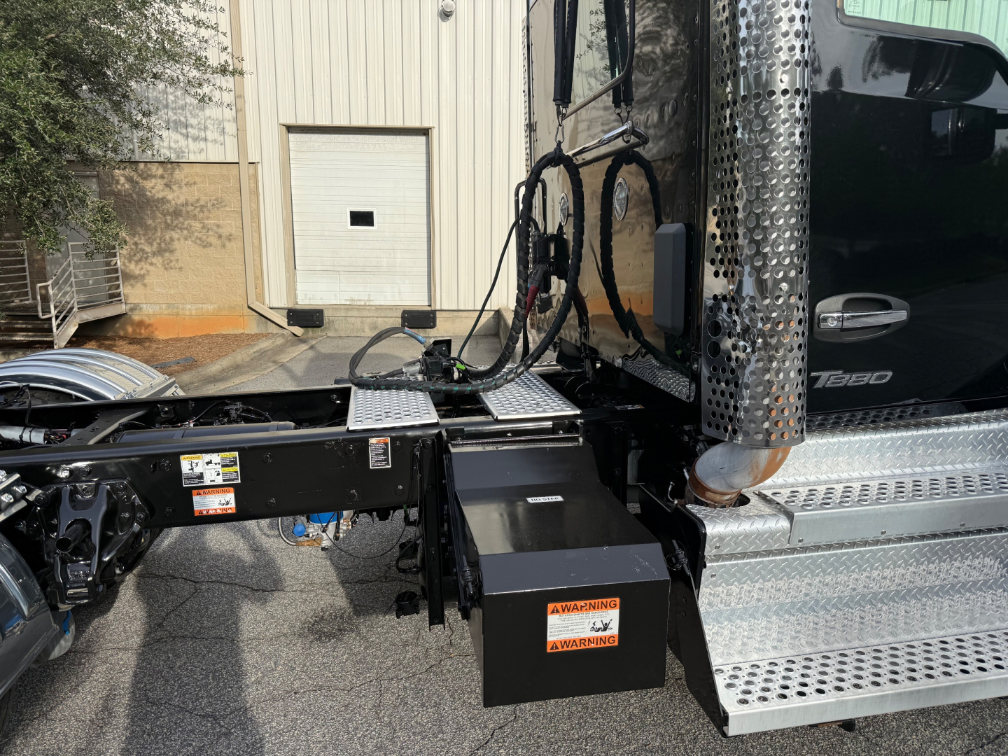 Kenworth T880 chassis and steps showing fuel/battery compartments and metallic chrome side steps; warning label on black toolbox; visible exhaust pipe and cab area.