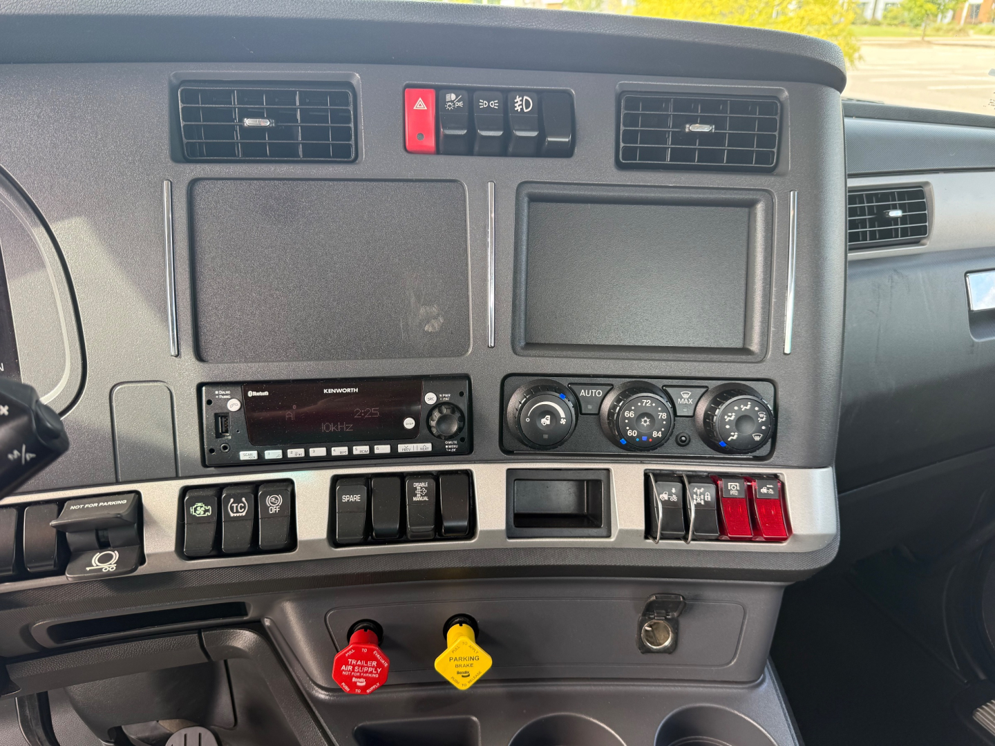 Kenworth T880 dashboard area showing dual vents, hazard switch, radio, climate controls, and multiple switches; red and yellow warning caps visible.