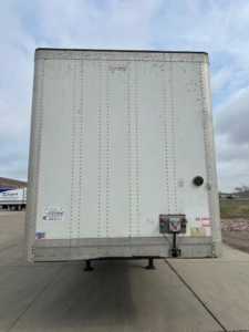 Trailer rear view showing vertical riveted doors and rear bumper with hitch, white exterior, small decals, and support legs visible.