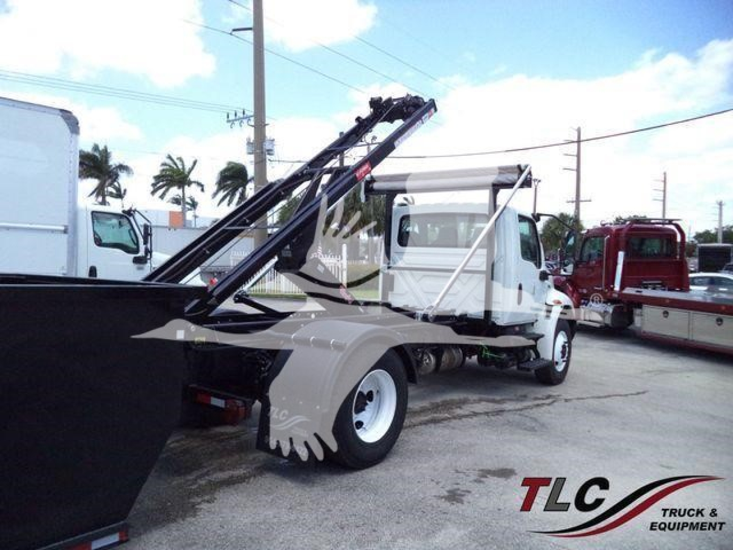 2019 International 4300 roll-off truck with a hydraulic transfer crane in the bed. Visible text includes U.S. VIN partially on windshield area and stock lot signage.
