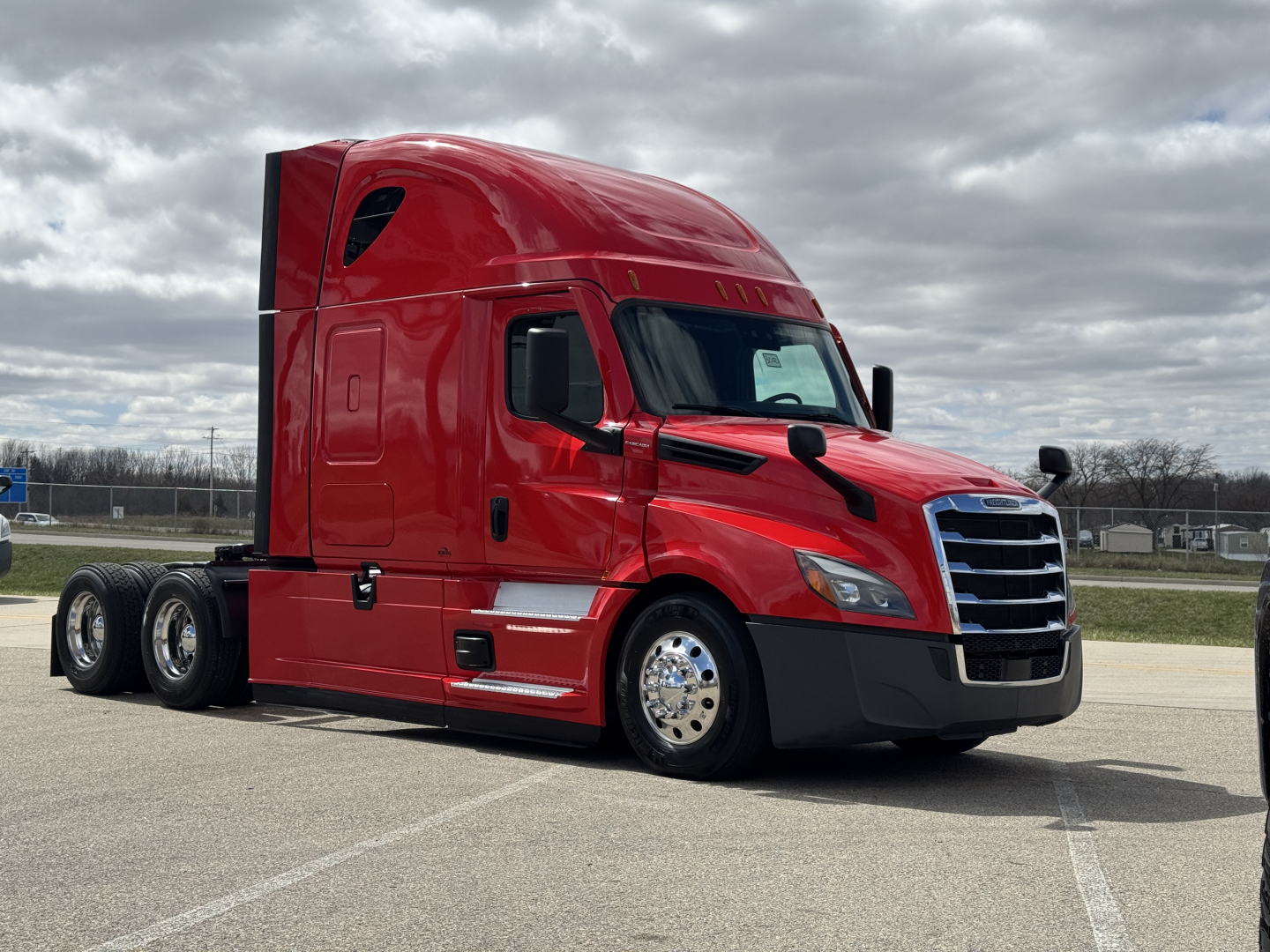 Freightliner Cascadia 126 red semi-truck in a parking lot, side view showing sleeper cab, chrome front grille, black bumper, dual rear axles.