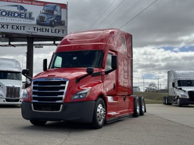 Red Freightliner Cascadia 126 semi-truck in a lot, front-left view with cab, chrome grille, black bumper, side mirrors, daytime overcast sky.