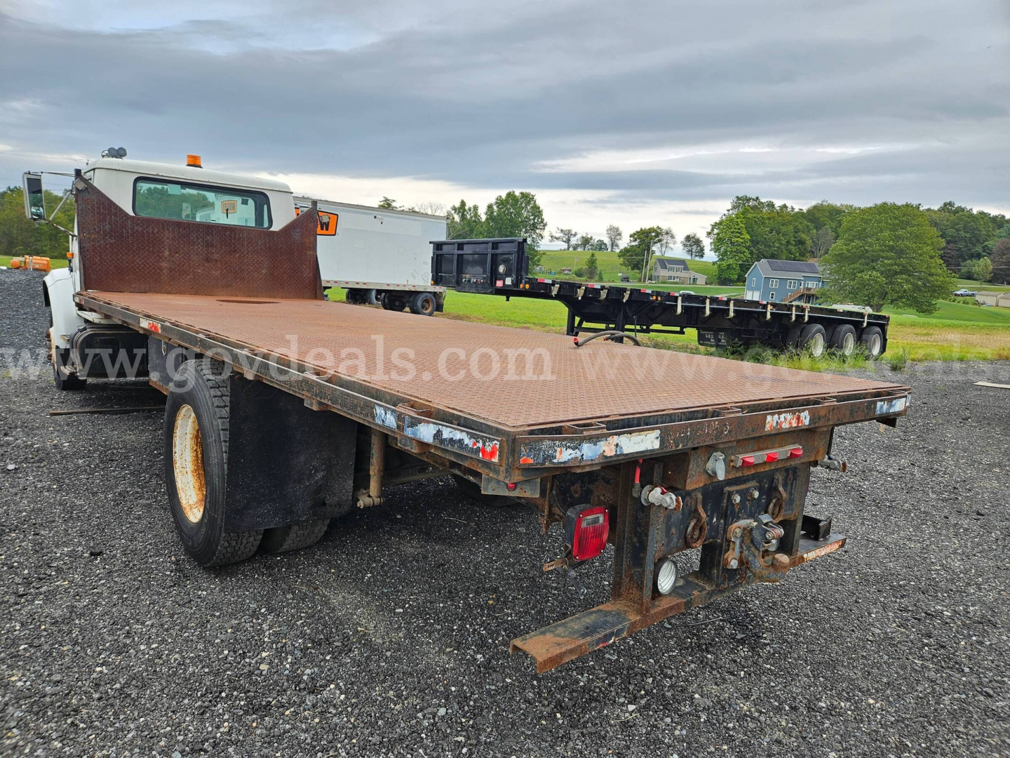 Flatbed dump truck rear angle showing steel flatbed with rust-free edges, dual rear axles, and hitch/detachable ramp area on a gravel lot.