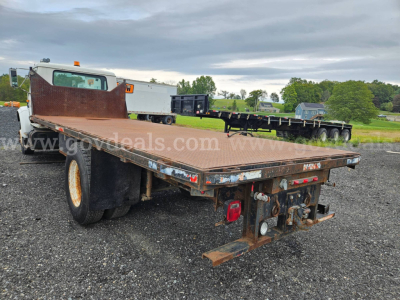 Flatbed dump truck rear angle showing steel flatbed with rust-free edges, dual rear axles, and hitch/detachable ramp area on a gravel lot.