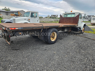 Flatbed dump truck chassis with long rusty flatbed and dual rear wheels, International 4900 style cab in background on a gravel lot.