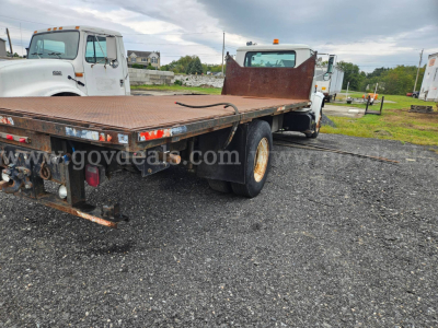 Flatbed dump truck rear and cargo deck showing steel flatbed with hydraulic hydraulics, dual rear wheels, and rear lights in a vehicle lot.