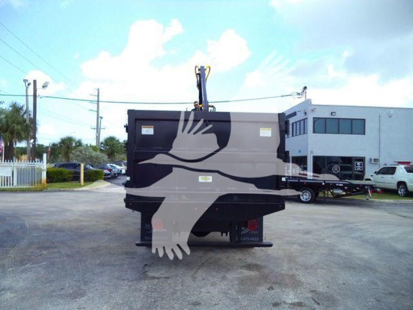 Rear view of a Ford F650 grapple truck with a crane and grapple attachment, logo of a bird on the hopper, visible dealership building and lot in background.