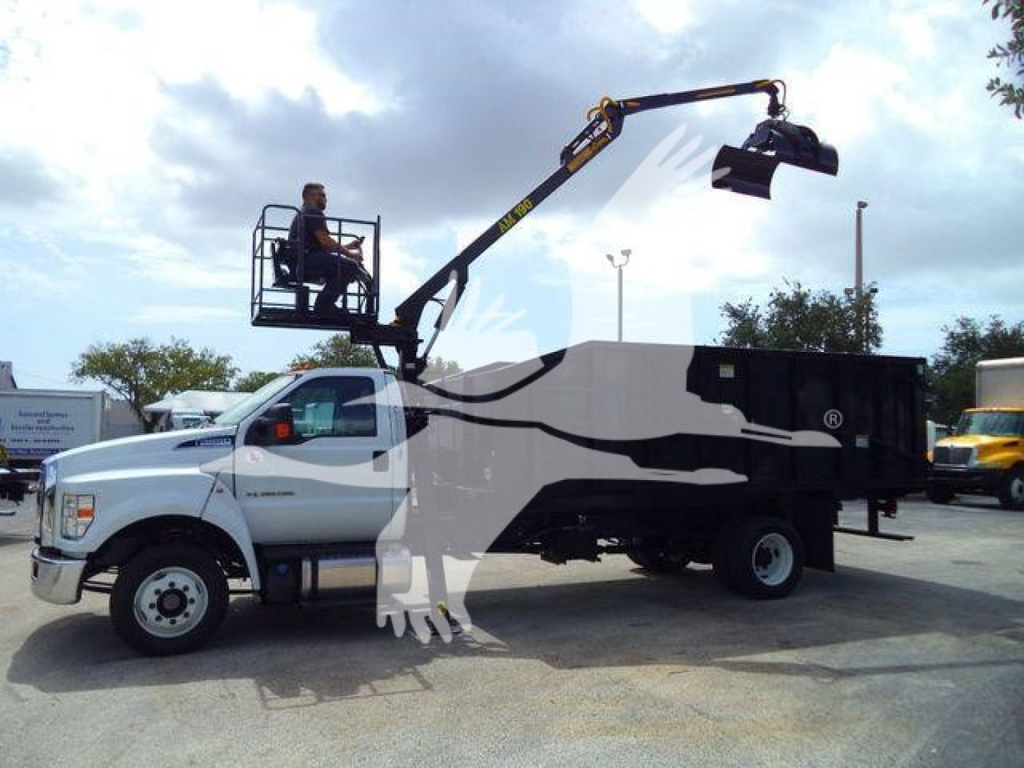 Ford F650 dump truck with grapple arm and operator in elevated basket performing loading operation.