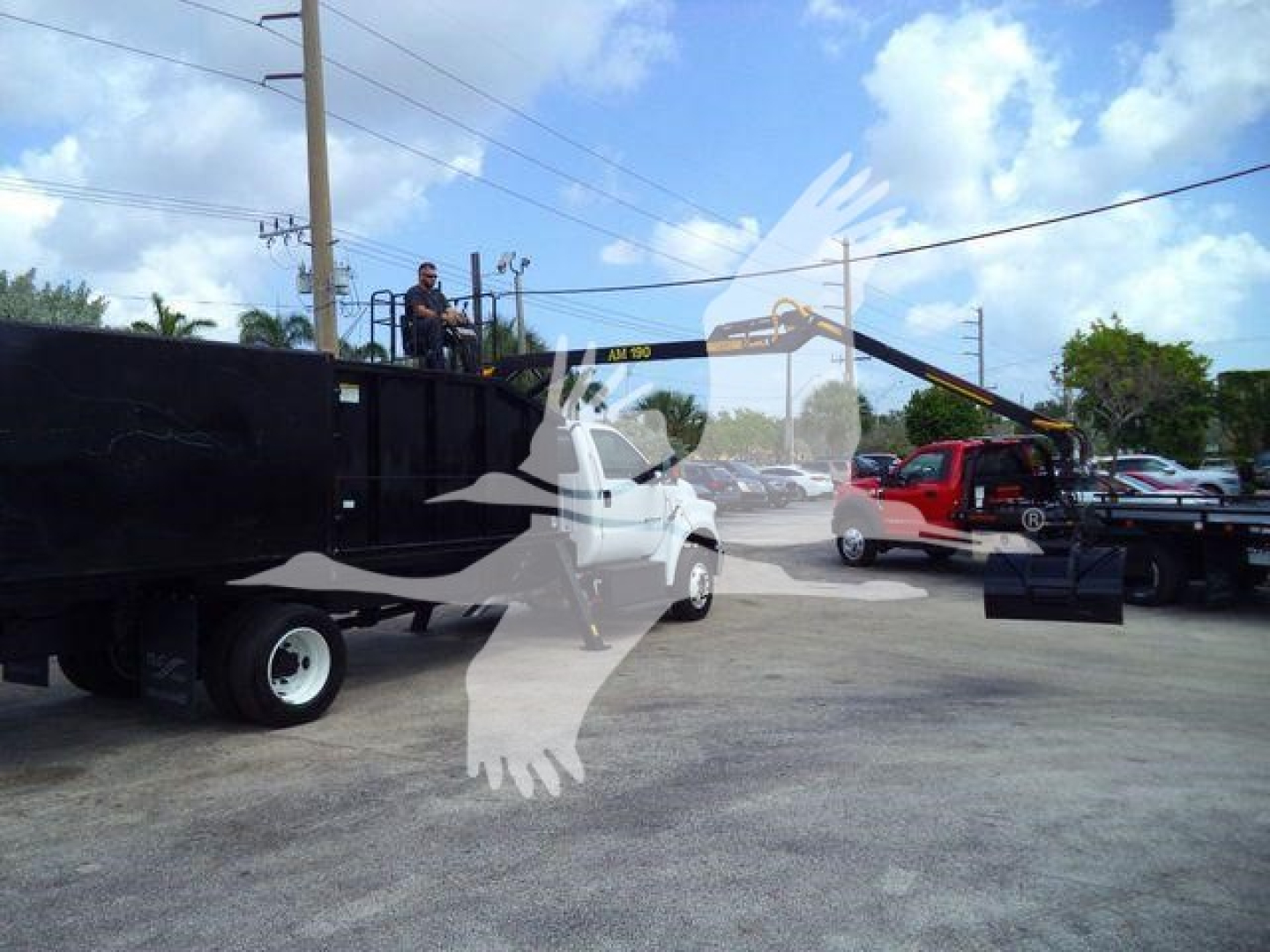 View of a Ford F-650 truck with a grapple arm attached on a trailer; branding and crane arm visible, with a red vehicle nearby.
